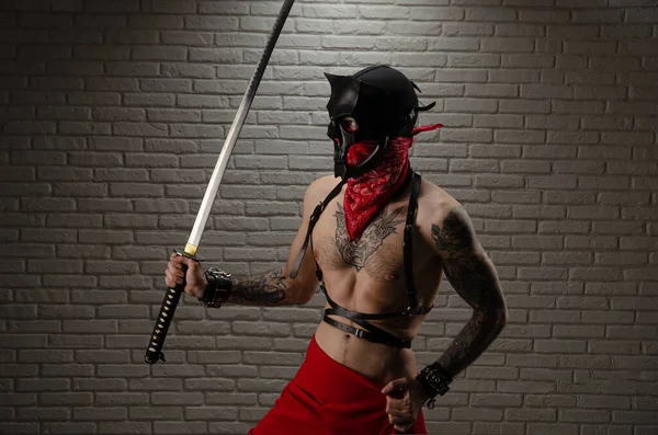 İblis maskeli bir samuray. Dövmeleri ve elinde tuğla bir duvarın arkasına dayanmış bir katana kılıcı var. — Stok fotoğraf