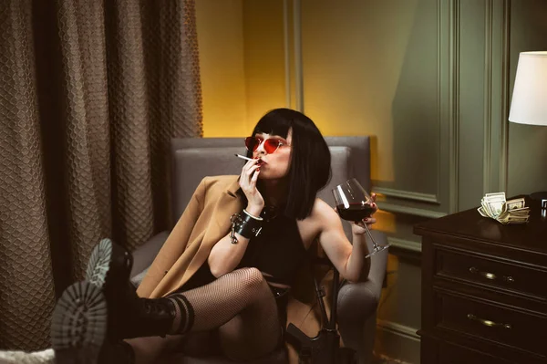 Uma criminosa em um hotel senta-se em uma cadeira fumando um cigarro e bebendo vinho — Fotografia de Stock