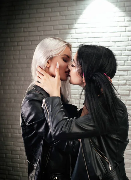 Två unga lesbiska lgbt kvinnor i läderjackor kyss på läpparna mot en vit tegelvägg — Stockfoto