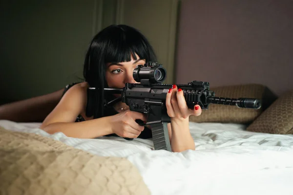 En kvinnlig lönnmördare på ett hotell ligger på en säng och håller ett automatgevär med en teleskopisk syn på offret medan begå mord — Stockfoto