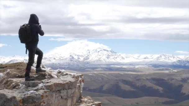 A male tourist traveler on a hill overlooking the mountain range of the Caucasus mountains Elbrus stands boldly on a rock and takes pictures on his phone — Stock Video