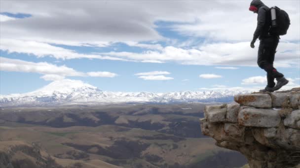 Ένας άνδρας τουρίστας ταξιδιώτης σε ένα λόφο με θέα την οροσειρά του Καυκάσου Όρη Elbrus στέκεται με τόλμη σε ένα γκρεμό — Αρχείο Βίντεο