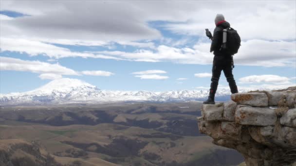 En manlig turist resenär på en kulle med utsikt över bergskedjan i Kaukasus bergen Elbrus står djärvt på en klippa och tar bilder på sin telefon — Stockvideo