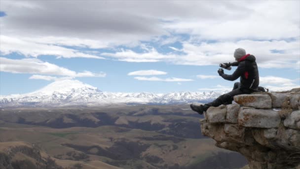 Ein männlicher Reisender auf einem Hügel mit Blick auf die Bergkette des Kaukasus Elbrus sitzt und trinkt Tee aus einer Thermoskanne — Stockvideo