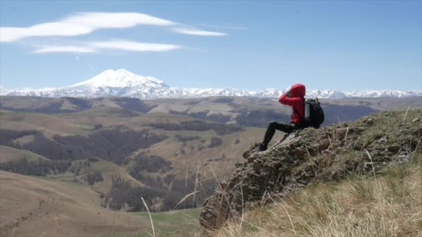 Ein männlicher Reisender auf einem Hügel mit Blick auf die Bergkette des Kaukasus Elbrus sitzt und trinkt Tee aus einer Thermoskanne — Stockvideo