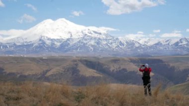 Kafkasya dağlarının sıradağlarına bakan bir erkek turist Elbrus cesurca bir kayanın üzerinde duruyor ve telefonuyla fotoğraf çekiyor.