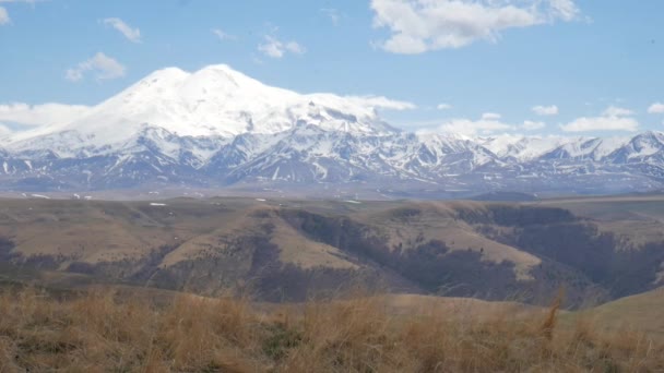 Un viajero turístico masculino en una colina con una vista de la cordillera de las montañas del Cáucaso Elbrus camina a lo largo de la ladera de la montaña contra — Vídeos de Stock