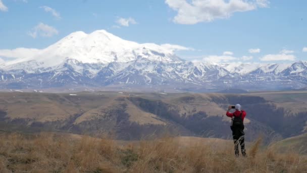 Ένας άνδρας τουρίστας ταξιδιώτης σε ένα λόφο με θέα την οροσειρά του Καυκάσου βουνά Elbrus στέκεται με τόλμη σε ένα βράχο και παίρνει φωτογραφίες στο τηλέφωνό του — Αρχείο Βίντεο