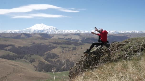 Чоловік - турист на пагорбі з видом на гірський хребет Кавказьких гір Ельбрус сміливо стоїть на скелі і фотографує свій телефон. — стокове відео