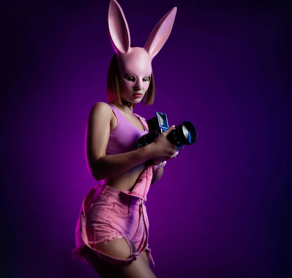 Stylish woman in pink short clothes and pink rabbit mask with retro video camera on neon background — Stock Photo, Image