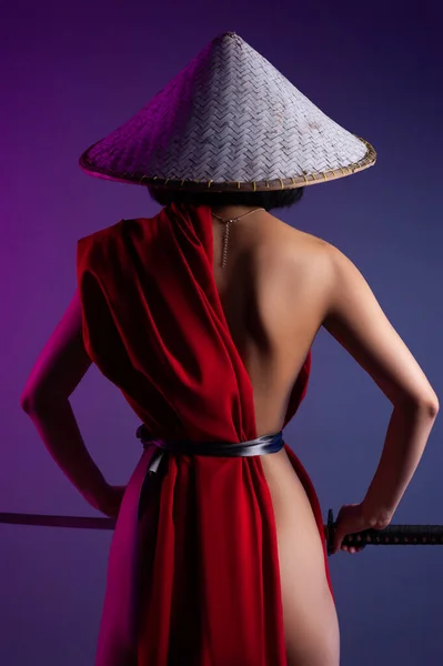 nude slender Asian woman in a red cape and an Asian hat with a katana in her hand image of a samurai on a neon background view from the back