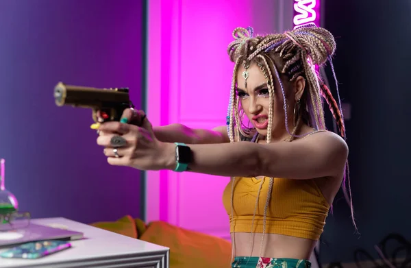 Una giovane donna hooligan con dreadlocks in abiti vivaci con una pistola esprime emozioni in luce al neon — Foto Stock