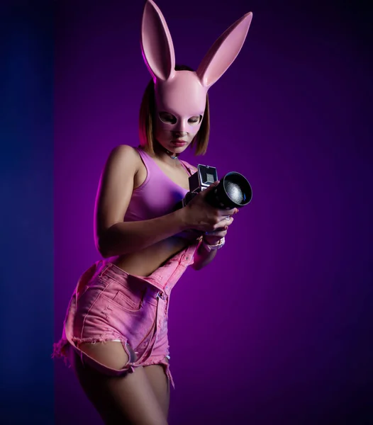 Mujer elegante en ropa corta rosa y máscara de conejo rosa con cámara de vídeo retro sobre fondo de neón —  Fotos de Stock