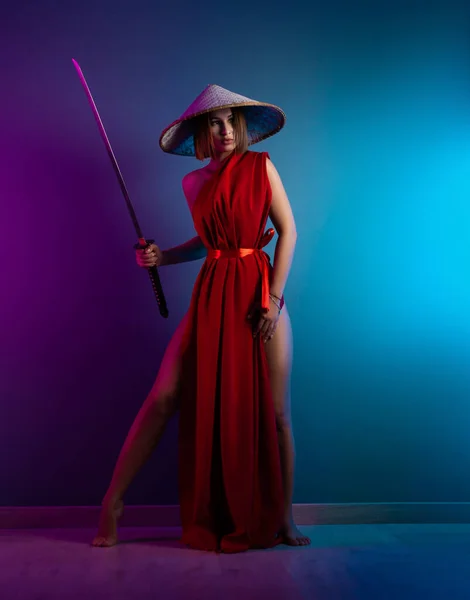 Sexy mujer en una capa roja y un sombrero asiático con una katana en la mano imagen de un samurai en color neón —  Fotos de Stock