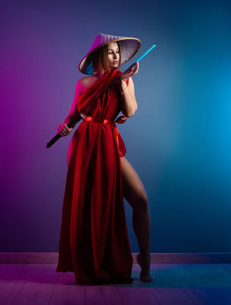 Mulher sexy em uma capa vermelha e um chapéu asiático com uma katana em sua imagem mão de um samurai na cor de néon — Fotografia de Stock