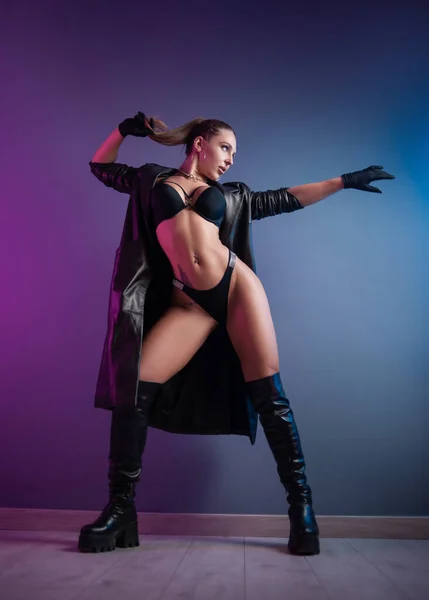 Donna sexy in biancheria intima, impermeabile in pelle e scarpe lunghe in pelle in studio su uno sfondo al neon — Foto Stock