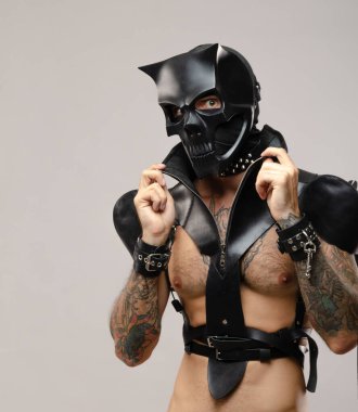 BDsm iblis kafatası maskeli bir adam deri bir pelerin giymiş ve vücudunda deri bilezikler ve kayışlar var.