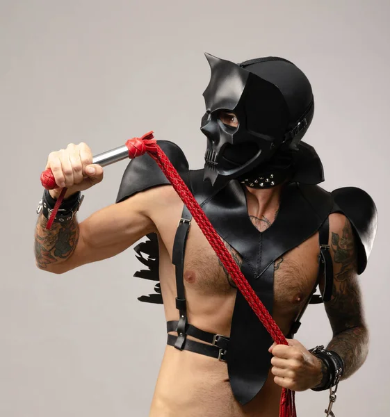 Egy férfi szado-mazo-ban koponya démon maszkot visel ostorral, bőresőkabátba öltözve bőrkarkötőkkel és szíjakkal a testén. — Stock Fotó