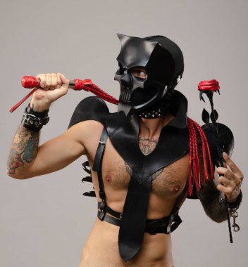 Kafatası iblis maskesi takan, kırbaçlı ve gül çiçekli deri yağmurluk giymiş deri bilezikler takan ve vücudunda kayışlar olan bir adam.