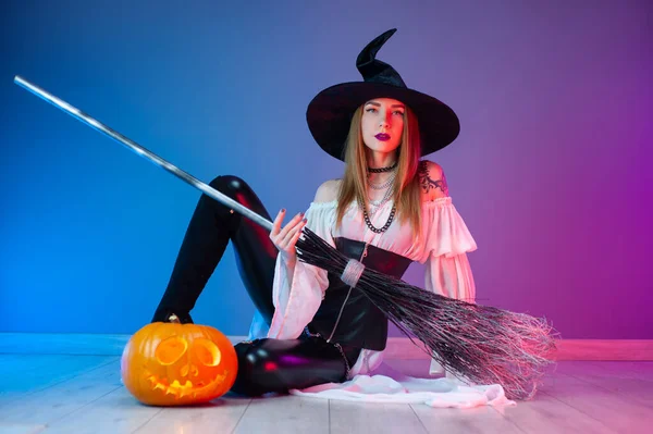 En flicka i häxdräkt för Halloween med kvast och en pumpa i neonljus — Stockfoto