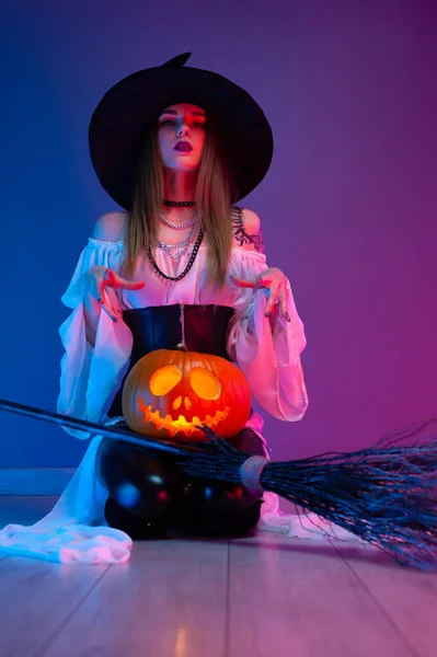 Uma menina em um traje de bruxa para o Halloween com uma vassoura e uma abóbora em luz de néon — Fotografia de Stock