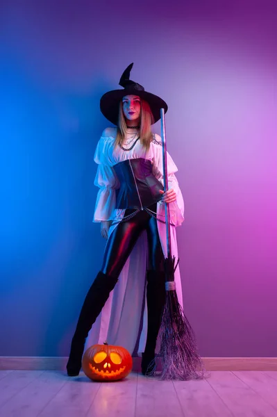 할로윈에 마녀 의상을 입고 빗자루를 들고 네온 라이트를 탄 호박을 입은 소녀 — 스톡 사진