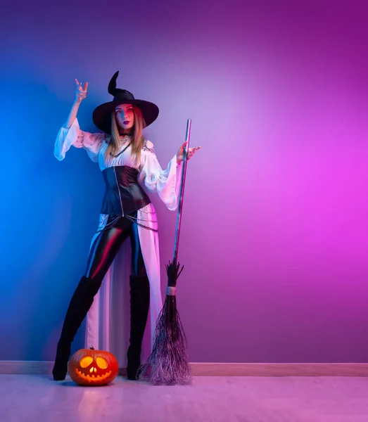 Dziewczyna w stroju czarownicy na Halloween z miotłą i dynią w neonowym świetle — Zdjęcie stockowe