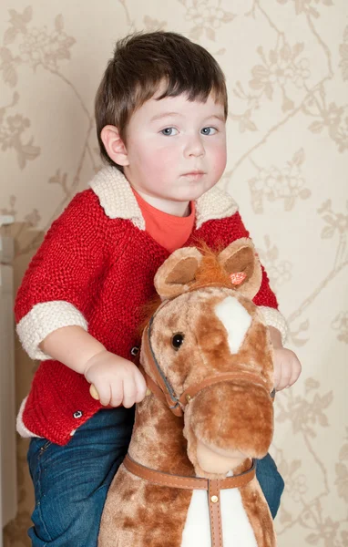 O rapaz senta-se em um brinquedo de um cavalo no quarto — Fotografia de Stock