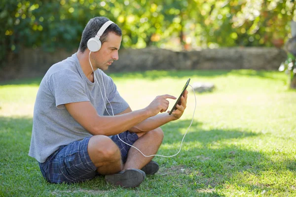 Giovane uomo che si rilassa con un tablet pc ascoltando musica con cuffie su un parco — Foto Stock