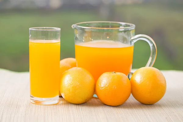 Стакан апельсинового сока и апельсинов на столе в саду — стоковое фото