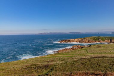 A Coruna 'da kıyı manzarası kayalık uçurumlar, dalgalar onlara çarpıyor ve açık mavi bir gökyüzü.