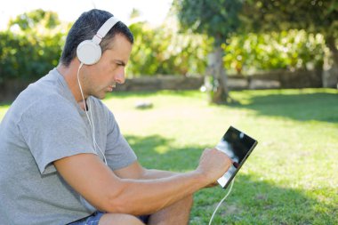 genç adam bir kulaklık ile müzik dinleme tablet pc ile rahatlatıcı bir parkta, açık