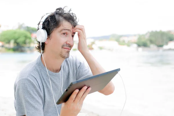 Young man holding a tablet with headphones, at the beach — Stock Photo, Image