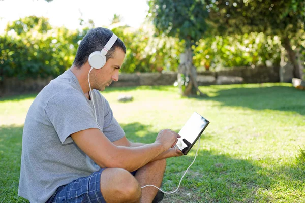 Giovane uomo che si rilassa con un tablet pc ascoltando musica con cuffie su un parco — Foto Stock