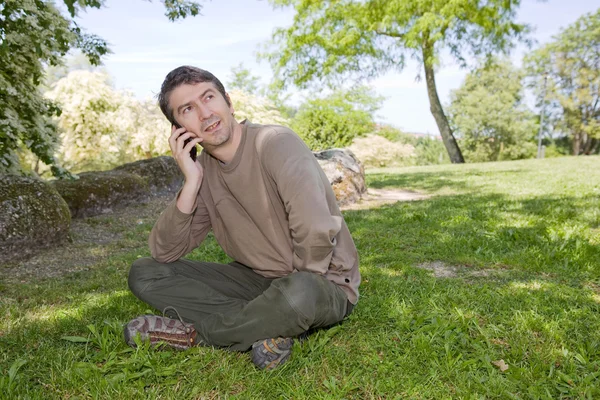 Young man sitting and talk on mobile phone, against green of summer park. — Stock Photo, Image