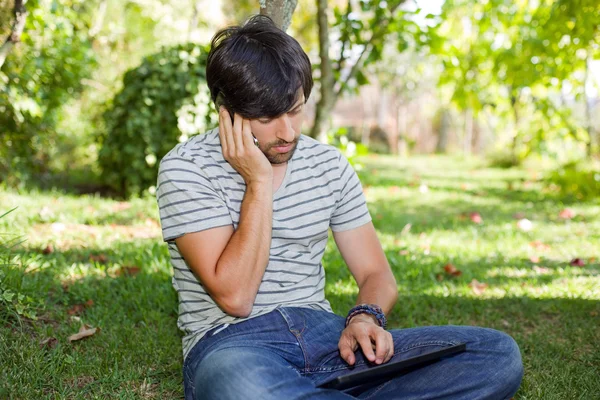 Νέος άνθρωπος χαλάρωση με ένα tablet pc και τηλέφωνο σε ένα πάρκο, υπαίθρια — Φωτογραφία Αρχείου