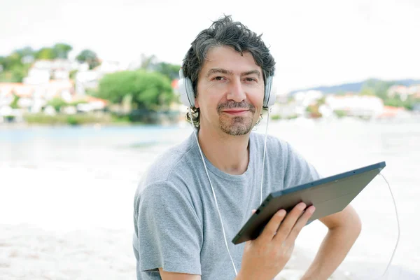 Ung man innehar en tablett med hörlurar, på stranden — Stockfoto