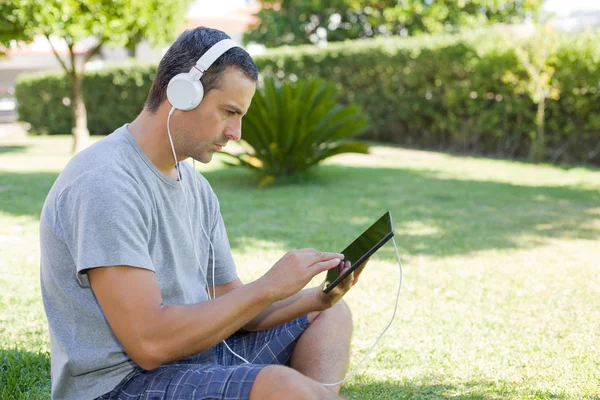 Junger Mann entspannt sich mit einem Tablet-PC und hört Musik mit Kopfhörern im Park — Stockfoto