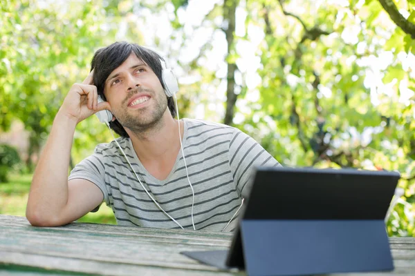 Mladý muž pracuje s tablet pc poslouchat hudbu se sluchátky na dřevěný stůl — Stock fotografie