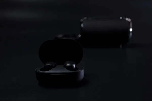 Black wireless headphones in charging case on black with wireless speaker on background.
