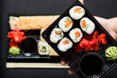 Hand holding set of sushi in plastic box close up, on black background.