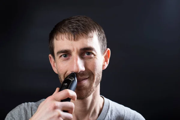 Portret of man using nose trimmer close up, on black background.