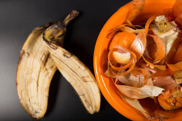 Banana peels and other peels from kitchen in orange plate, on black background, close up.