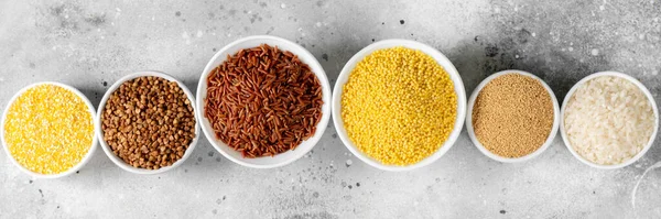 White and red rice, buckwheat, amaranth seeds, corn groats, quinoa and millet in white bowls on the light gray kitchen table. Gluten-free cereals. Top view