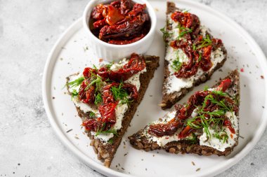 Krem peynir ve güneşte kurutulmuş domates. Kepekli ekmek, krem peynir ve güneşte kurutulmuş domateslerden yapılmış sandviç ya da crostini. Açık gri mutfak masasında beyaz bir tabakta