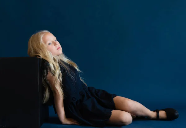 beautiful blonde in a dark blue dress on a solid blue background. The photo is great for demonstrating the character of a child - daydreaming, enthusiasm, vulnerability, touching, beauty, stubbornness, harm, cunning, coquetry.