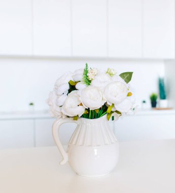 Yumuşak ev dekorasyonu, beyaz sürahi, beyaz duvarda beyaz güzel çiçekler olan vazo ve ahşap bir rafta. İç mekan.