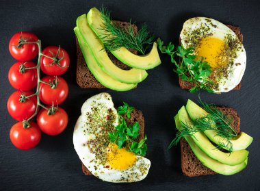 Tam tahıllı sandviç, kızarmış bıldırcın yumurtası, avokado, otlar ve siyah arka planda tohumlar.