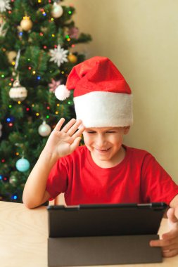 Kırmızı Noel şapkalı şirin çocuk tablet kullanarak video çağrısı yapıyor.