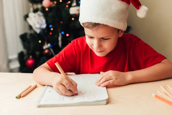 Glimlachend kind jongen schrijven brief aan de Kerstman, kerstboom op de achtergrond — Stockfoto